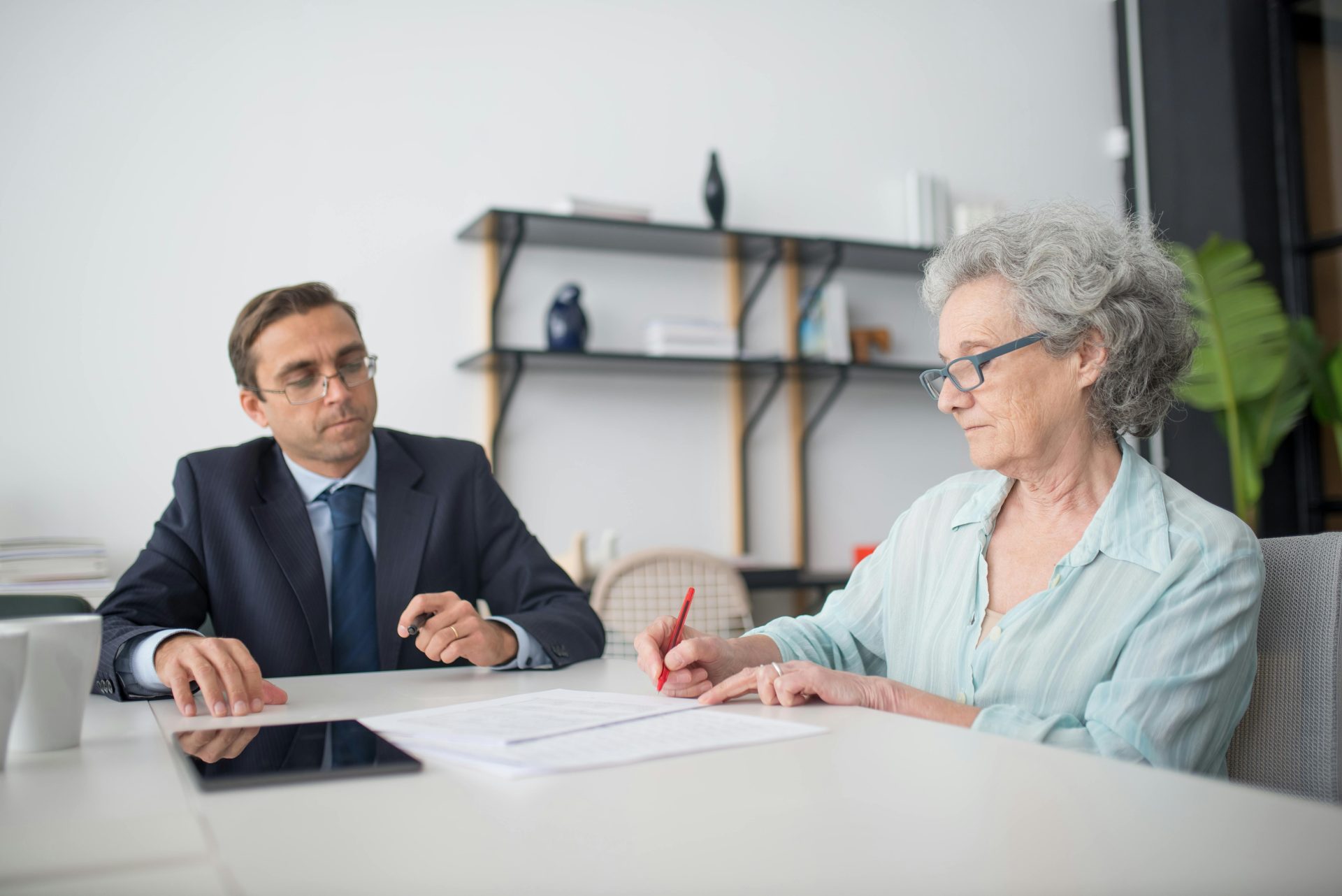 Une femme âgée signe des documents dans un bureau moderne en présence d'un consultant.